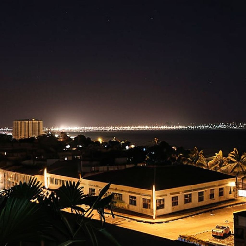 ** A nighttime scene in Maputo, Mozambique. The city is mostly dark, with only a few scattered lights visible in the distance. Focus on a hotel room with a window open to show the darkness and distant lights. Emphasize the feeling of heat and the sounds of the city.
**